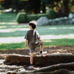 Young boy pictured from behind, standing on the bank of a river, leaning on a large tree root.